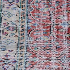 May include: Close-up of a section of a vintage rug with a distressed, faded appearance. The rug features a complex pattern in shades of red, beige, blue, and green. The design includes floral and geometric elements, with a black border.