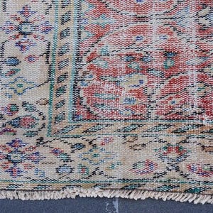 May include: Close-up of a section of a vintage rug with a distressed appearance. The rug features a central field of red and cream hues, with intricate floral patterns in blue, green, and black. The border is cream with floral motifs and a fringed edge.