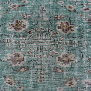 May include: Close-up of a distressed, vintage rug with a teal and brown floral pattern. The rug features a central medallion design and faded details, giving it an antique appearance. The rug's texture is visible, showcasing its woven construction.