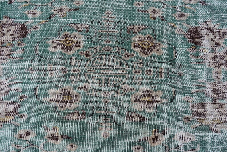 May include: Close-up of a distressed, vintage rug with a teal and brown floral pattern. The rug features a central medallion design and faded details, giving it an antique appearance. The rug's texture is visible, showcasing its woven construction.