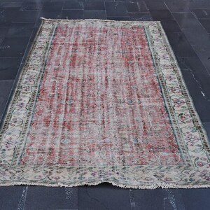 May include: Rectangular, distressed rug with a faded red and cream central field. The rug features a detailed border with floral and geometric patterns in shades of green, blue, and cream. The rug is placed on a dark gray tiled floor.