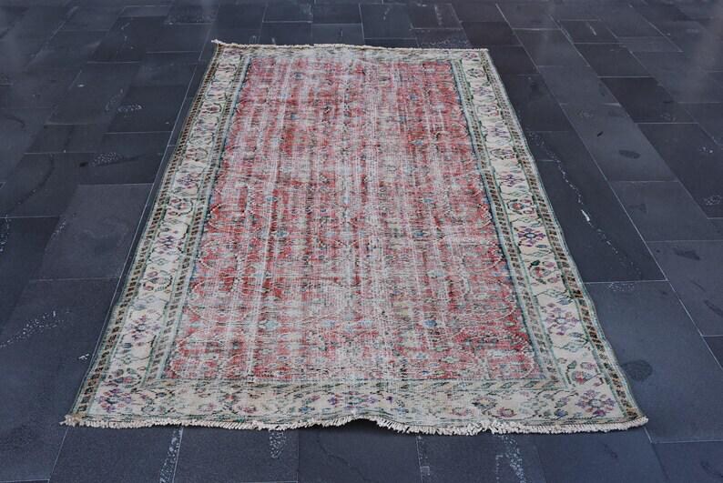 May include: Rectangular, distressed rug with a faded red and cream central field. The rug features a detailed border with floral and geometric patterns in shades of green, blue, and cream. The rug is placed on a dark gray tiled floor.