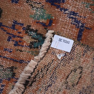 May include: Close-up of a brown and beige rug with a blue, green, and black geometric pattern. A white tag with the text "DC 11265" is attached to the rug.