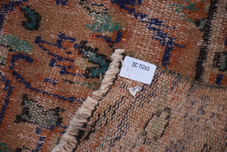 May include: Close-up of a brown and beige rug with a blue, green, and black geometric pattern. A white tag with the text "DC 11265" is attached to the rug.