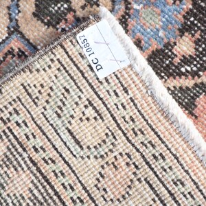May include: Close-up of the back of a rug with a white fringe. The rug has a woven pattern of brown, beige, and light pink. A white tag with the text "DC10851" is attached to the rug.