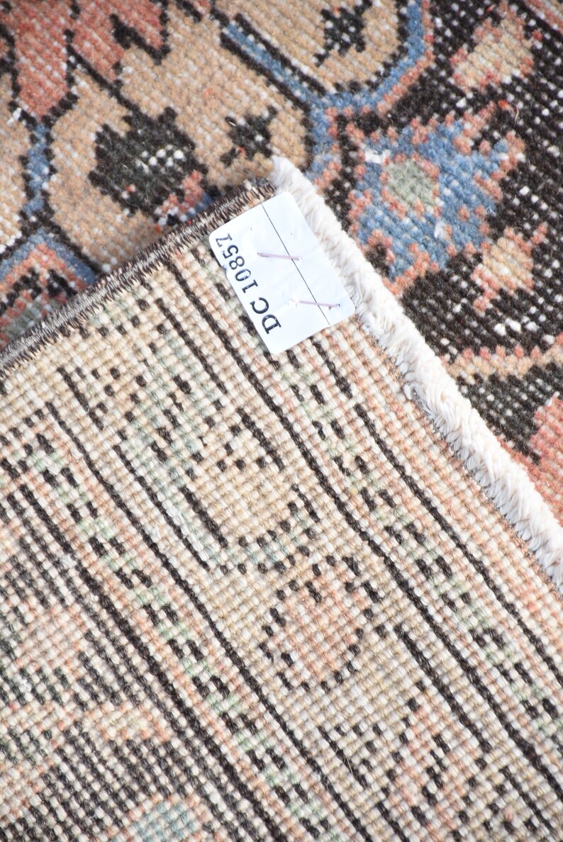 May include: Close-up of the back of a rug with a white fringe. The rug has a woven pattern of brown, beige, and light pink. A white tag with the text "DC10851" is attached to the rug.