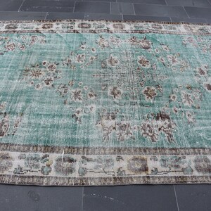 May include: A large, rectangular rug with a distressed, vintage appearance. The rug features a central field of teal green with faded floral patterns in brown and cream. A cream border with floral and geometric designs frames the rug.