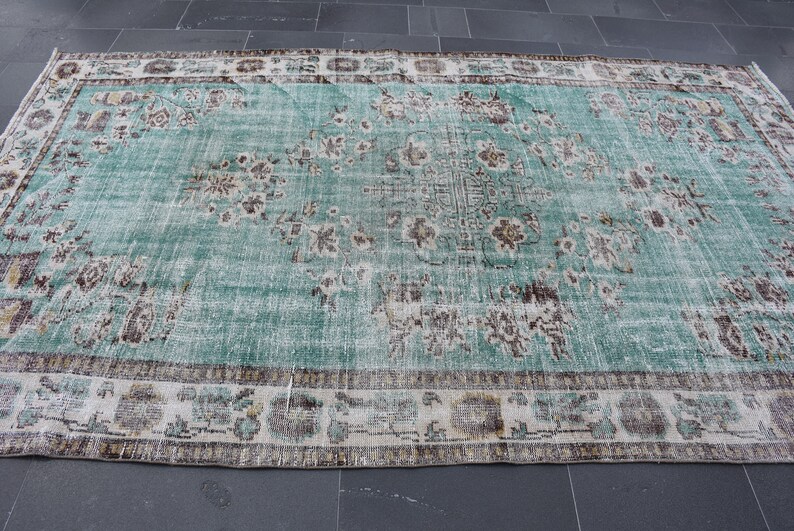 May include: A large, rectangular rug with a distressed, vintage appearance. The rug features a central field of teal green with faded floral patterns in brown and cream. A cream border with floral and geometric designs frames the rug.
