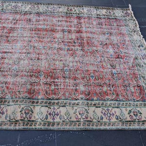 May include: A rectangular, distressed rug with a faded red and cream central field. The rug features a detailed border with floral and geometric patterns in shades of cream, green, and purple. The rug is placed on a dark surface.