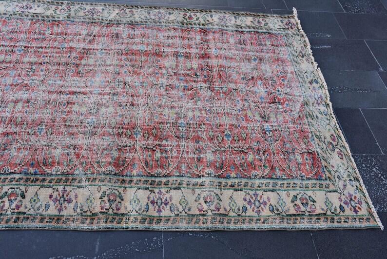 May include: A rectangular, distressed rug with a faded red and cream central field. The rug features a detailed border with floral and geometric patterns in shades of cream, green, and purple. The rug is placed on a dark surface.