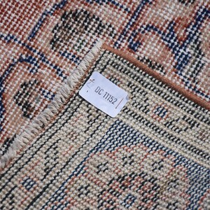 May include: Close-up of a section of a vintage rug with a detailed pattern. The rug features a combination of beige, brown, and navy blue colours. The tag reads "DC 11152". The rug has a fringed edge.
