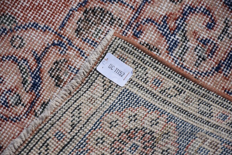 May include: Close-up of a section of a vintage rug with a detailed pattern. The rug features a combination of beige, brown, and navy blue colours. The tag reads "DC 11152". The rug has a fringed edge.