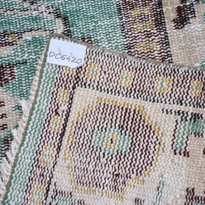 May include: Close-up of a section of a vintage rug with a distressed look. The rug features a cream-colored background with a repeating pattern in shades of brown, green, and yellow. The tag in the image reads "DC5420".