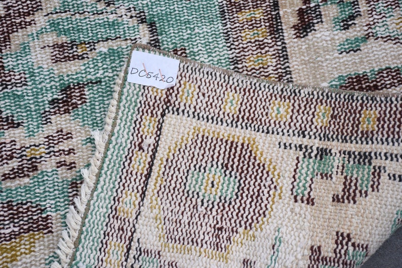 May include: Close-up of a section of a vintage rug with a distressed look. The rug features a cream-colored background with a repeating pattern in shades of brown, green, and yellow. The tag in the image reads "DC5420".