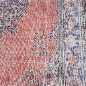 May include: Close-up of a vintage rug with a distressed, faded appearance. The rug features a central field of coral-red with intricate floral and geometric patterns in navy blue and cream. The rug's border is a darker shade of blue.