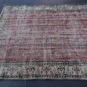 May include: A rectangular, distressed rug with a faded red and cream color palette. The rug features a central field of red with a repeating pattern and a cream-colored border with floral and geometric designs. The rug is placed on a dark surface.