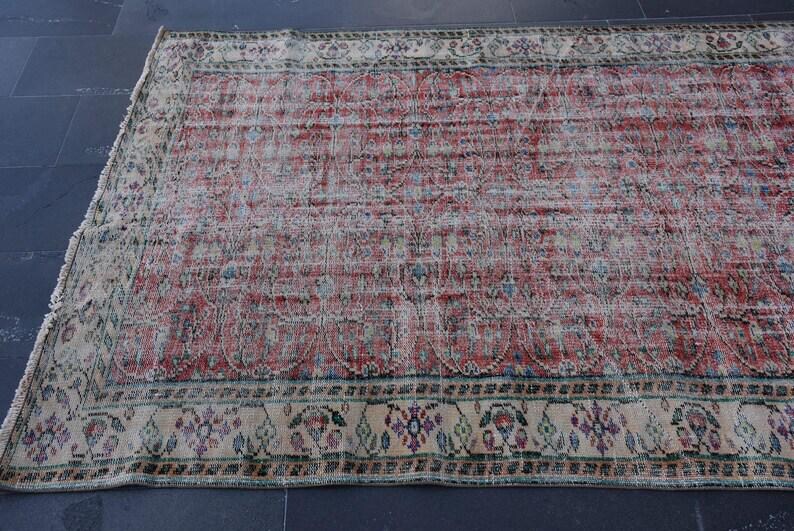 May include: A rectangular, distressed rug with a faded red and cream color palette. The rug features a central field of red with a repeating pattern and a cream-colored border with floral and geometric designs. The rug is placed on a dark surface.