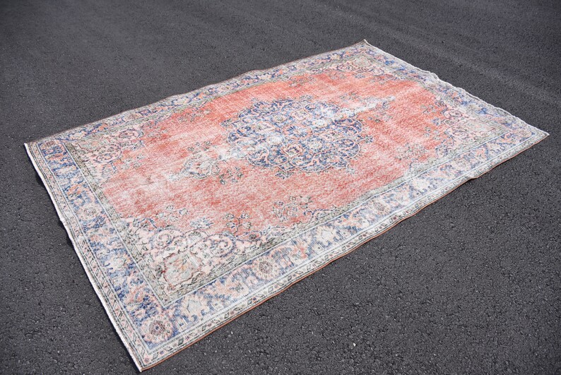 May include: A rectangular rug with a distressed appearance. It features a faded red central area and detailed blue and cream floral patterns. The rug has a border with similar blue and cream designs. It is placed on a dark surface.
