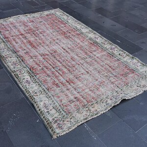 May include: A rectangular, distressed rug with a faded red and cream color palette. The rug features a central field of red with a floral border in cream, green, and purple. Frayed edges add to the vintage aesthetic.