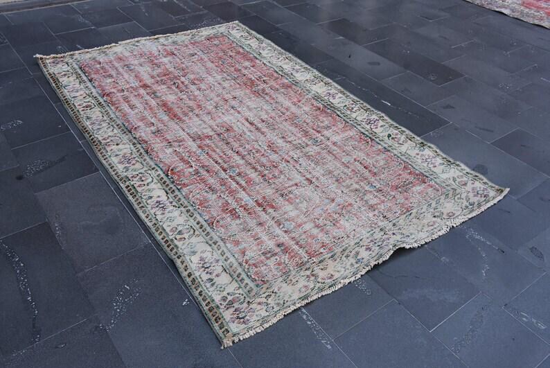 May include: A rectangular, distressed rug with a faded red and cream color palette. The rug features a central field of red with a floral border in cream, green, and purple. Frayed edges add to the vintage aesthetic.