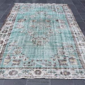 May include: A rectangular, distressed-look rug with a faded turquoise central field and a detailed border. The rug features floral and geometric patterns in shades of brown, beige, and white. The rug's dimensions are not visible in the image.