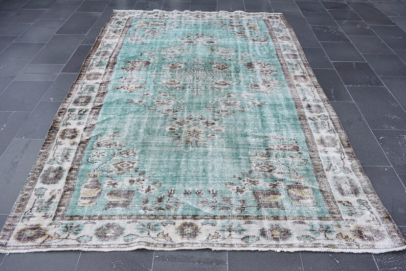 May include: A rectangular, distressed-look rug with a faded turquoise central field and a detailed border. The rug features floral and geometric patterns in shades of brown, beige, and white. The rug's dimensions are not visible in the image.