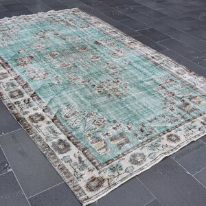 May include: A rectangular, distressed-look rug with a faded turquoise central field and a detailed border. The rug features floral and geometric patterns in shades of brown, beige, and white, creating a vintage aesthetic. The rug is suitable for home decor.