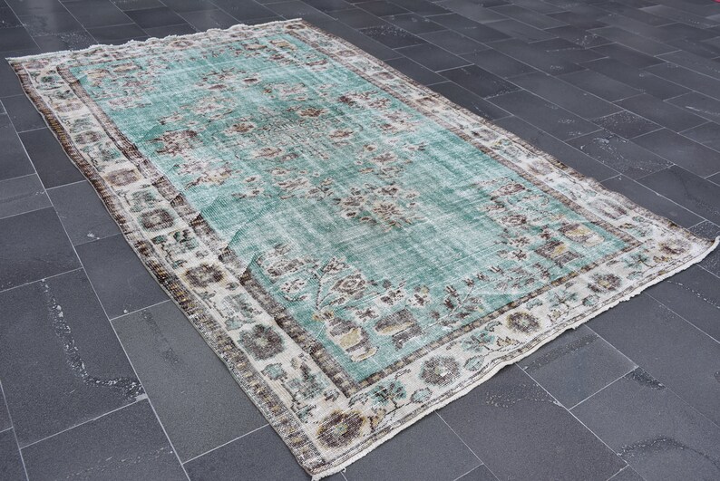 May include: A rectangular, distressed-look rug with a faded turquoise central field and a detailed border. The rug features floral and geometric patterns in shades of brown, beige, and white, creating a vintage aesthetic. The rug is suitable for home decor.