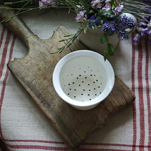 Op de afbeelding: Een witte keramische theezeef met zwarte perforaties ligt op een rustieke houten snijplank. Een kleine vaas met paarse en roze bloemen en groen staat op de achtergrond, met een rood-wit gestreepte doek.