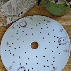 Può includere: Un piatto scolapiatti rotondo in porcellana bianca dotato di un foro centrale circolare e numerose piccole perforazioni sulla superficie. Il piatto è decorato con delicate ghirlande floreali viola e dettagli di fogliame verde dipinti a mano.