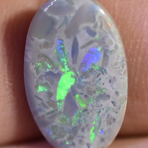 May include: Oval-shaped opal gemstone with a milky white base and vibrant flashes of color. The stone displays a play of color with green, blue, and purple hues. The opal's surface has a textured appearance, showcasing its natural beauty.