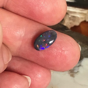 Pode incluir: Uma gema de opala ovalada de cor cinza escura com flashes de azul, verde e vermelho.