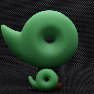 May include: Two green, abstract, spiral-shaped sculptures. The larger sculpture is the primary focus, with a smaller version of the same design below it. The sculptures are a solid, matte green color and sit on a dark background.