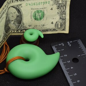 May include: A light green pendant with a spiral design, approximately 3 inches long, on a gold-colored cord. A smaller, matching spiral sits atop a wooden base. A one-dollar bill and a ruler are also in the image.