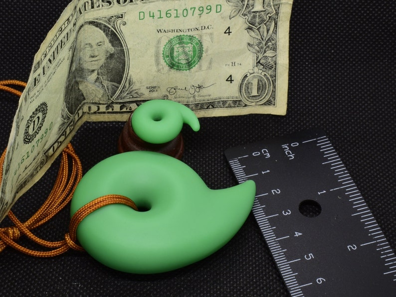 May include: A light green pendant with a spiral design, approximately 3 inches long, on a gold-colored cord. A smaller, matching spiral sits atop a wooden base. A one-dollar bill and a ruler are also in the image.