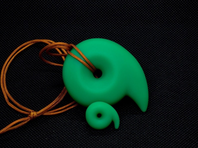 May include: A bright green pendant and smaller charm in a spiral design, with a matching cord. The pendant has a smooth, matte finish. The cord is a golden-brown color. The background is black.