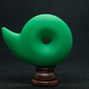 May include: A vibrant green, abstract sculpture with a spiral design, mounted on a dark wooden base. The sculpture has a smooth, matte finish and a circular opening in the center. The base is a dark brown color.