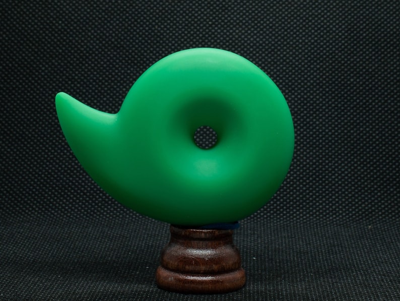 May include: A vibrant green, abstract sculpture with a spiral design, mounted on a dark wooden base. The sculpture has a smooth, matte finish and a circular opening in the center. The base is a dark brown color.