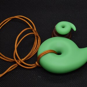 May include: A green pendant with a spiral design, strung on a gold-colored cord. The pendant has a smooth, matte finish and a small, matching spiral accent. The cord is knotted through the pendant's center.