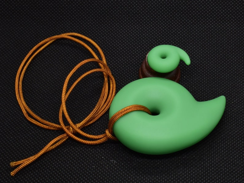 May include: A green pendant with a spiral design, strung on a gold-colored cord. The pendant has a smooth, matte finish and a small, matching spiral accent. The cord is knotted through the pendant's center.