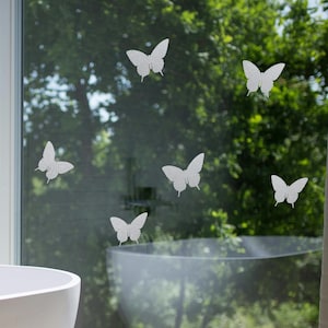 Könnte beinhalten: Eine moderne weiße Badewanne in einem Badezimmer. Die ovale Wanne steht auf einem hellen Boden. Weiße Schmetterlingsaufkleber zieren das Fenster mit Blick auf grüne Bäume. Die Wände sind aus weißen Ziegeln.