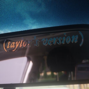 Puede incluir: Un espejo retrovisor con una pegatina que dice "(taylor's version)" en un degradado azul y naranja.