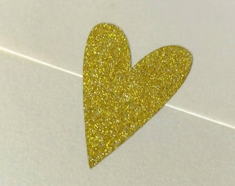 Puede incluir: Un sobre blanco con una pegatina de corazón de purpurina dorada.