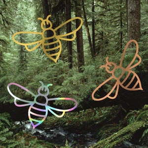May include: Three stylized bee illustrations in gold, copper, and iridescent finishes are set against a backdrop of a lush forest. The bees are depicted with detailed wings and segmented bodies, creating a nature-inspired design.