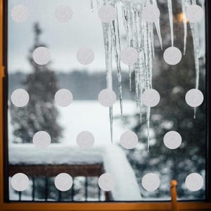 Op de afbeelding: Een raam versierd met zilveren glitter stipstickers. Door het glas is een besneeuwd landschap en ijspegels te zien. De stickers zijn in een patroon gerangschikt en geven de winterse scène een decoratieve uitstraling.
