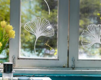 May include: A window with white ginkgo leaf decals. A cup of coffee and a small white cup with a spoon sit on a table. Yellow tulips are in a vase to the left of the window.