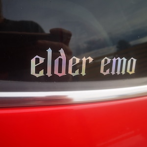 May include: A red car with a black window that has a silver iridescent sticker that says "elder emo" in a gothic font.