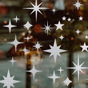 Puede incluir: Una colección de calcomanías plateadas con forma de estrella brillante de varios tamaños. Las estrellas están dispuestas en un patrón aleatorio, con algunas superpuestas. El fondo está borroso, lo que sugiere una ventana o superficie de vidrio. Las estrellas tienen una apariencia texturizada y brillante.
