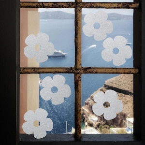 Może przedstawiać: Okno z rdzawobrązową metalową kratą, z pięcioma białymi, błyszczącymi naklejkami w kształcie kwiatów. Tło przedstawia malowniczy widok na morze, statek wycieczkowy i skaliste wybrzeże pod jasnym niebem.