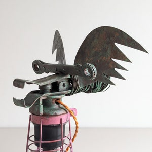 May include: A steampunk-style lamp with a pink base and a wire cage shade. The lamp is shaped like a bird with wings made of rusty metal.